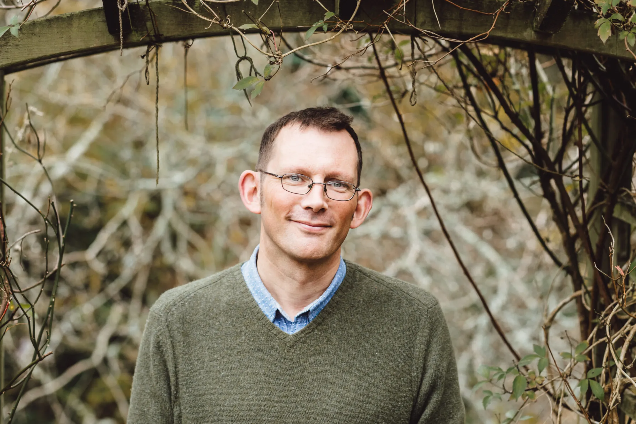 Photo of writer Rob Hopkins. He is wearing a green sweater, blue shirt and glasses. He is stood outside surrounded by a background of trees.